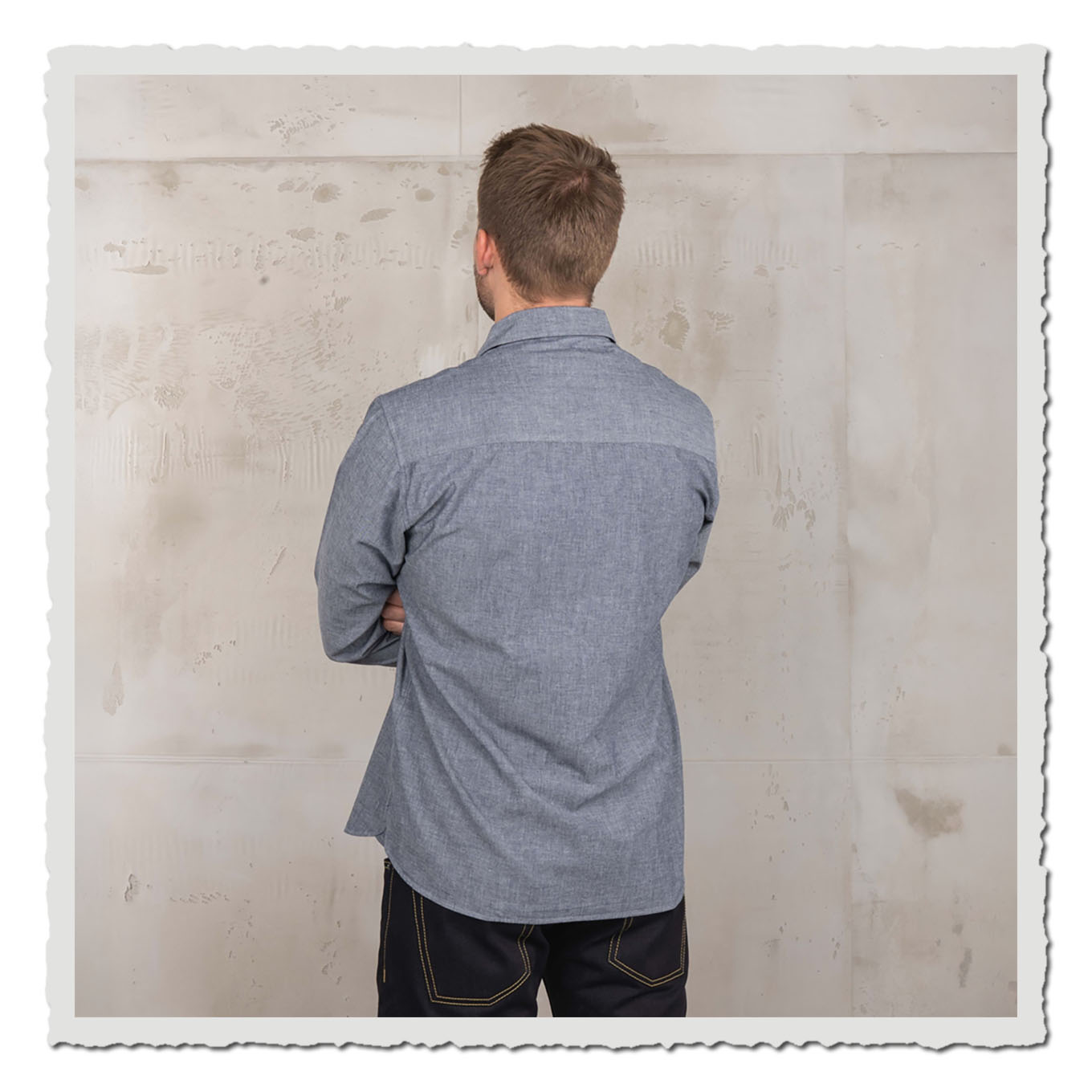 Rückansicht eines Models im hellblauen Langarmhemd vor heller, strukturierter Wand im Studio.